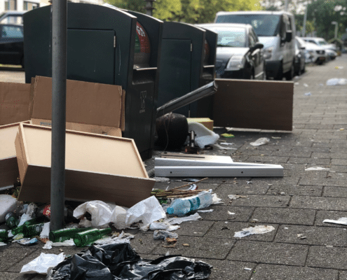 Foto van een afvalcontainer op straat. Naast de afvalcontainer liggen lege flessen, kapotte meubels en zwerfafval.
