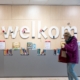 Foto genomen in Bibliotheek Segbroek. Een vrouw in een paarse jas loopt voor een wand met boeken langs. Op de wand staat 'welkom'.