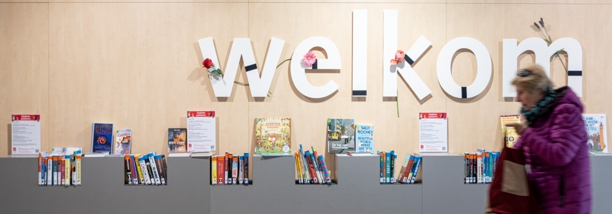 Foto genomen in Bibliotheek Segbroek. Een vrouw in een paarse jas loopt voor een wand met boeken langs. Op de wand staat 'welkom'.